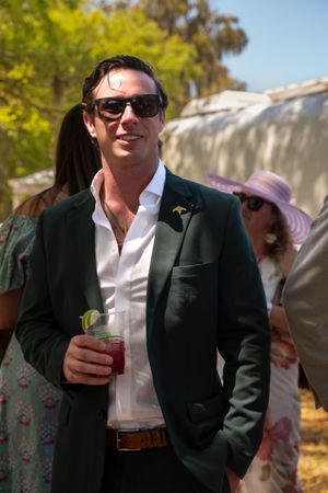 Man in dark green blazer and sunglasses holding a cocktail at an outdoor event.