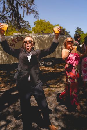 Man in suit raises drinks celebrating outdoors at a wedding reception with guests in floral dress nearby.