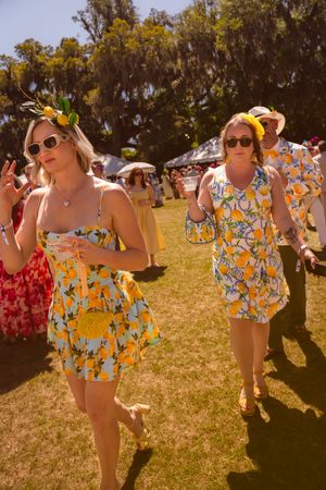 Two women wearing matching yellow lemon print outfits and sunglasses walk at an outdoor summer festival.