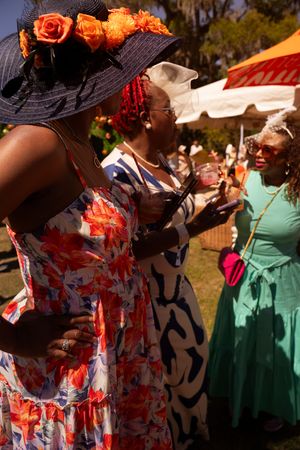 Three women in colorful floral dresses and decorative hats socialize outdoors at a sunny garden party event.