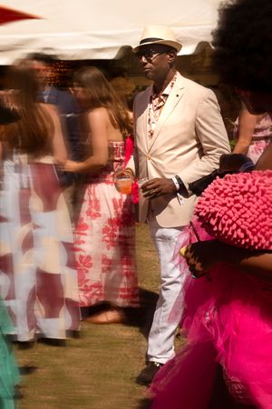 Stylish man in white suit and fedora standing at lively outdoor garden party event.
