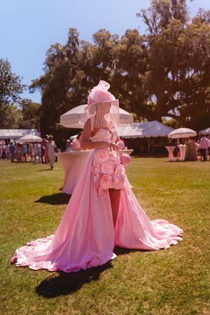 Person in elaborate pink gown with floral details and ornate hat stands on lawn at outdoor garden party event.