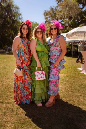 Three women pose in vibrant colorful dresses and pink floral fascinators at an outdoor garden party.