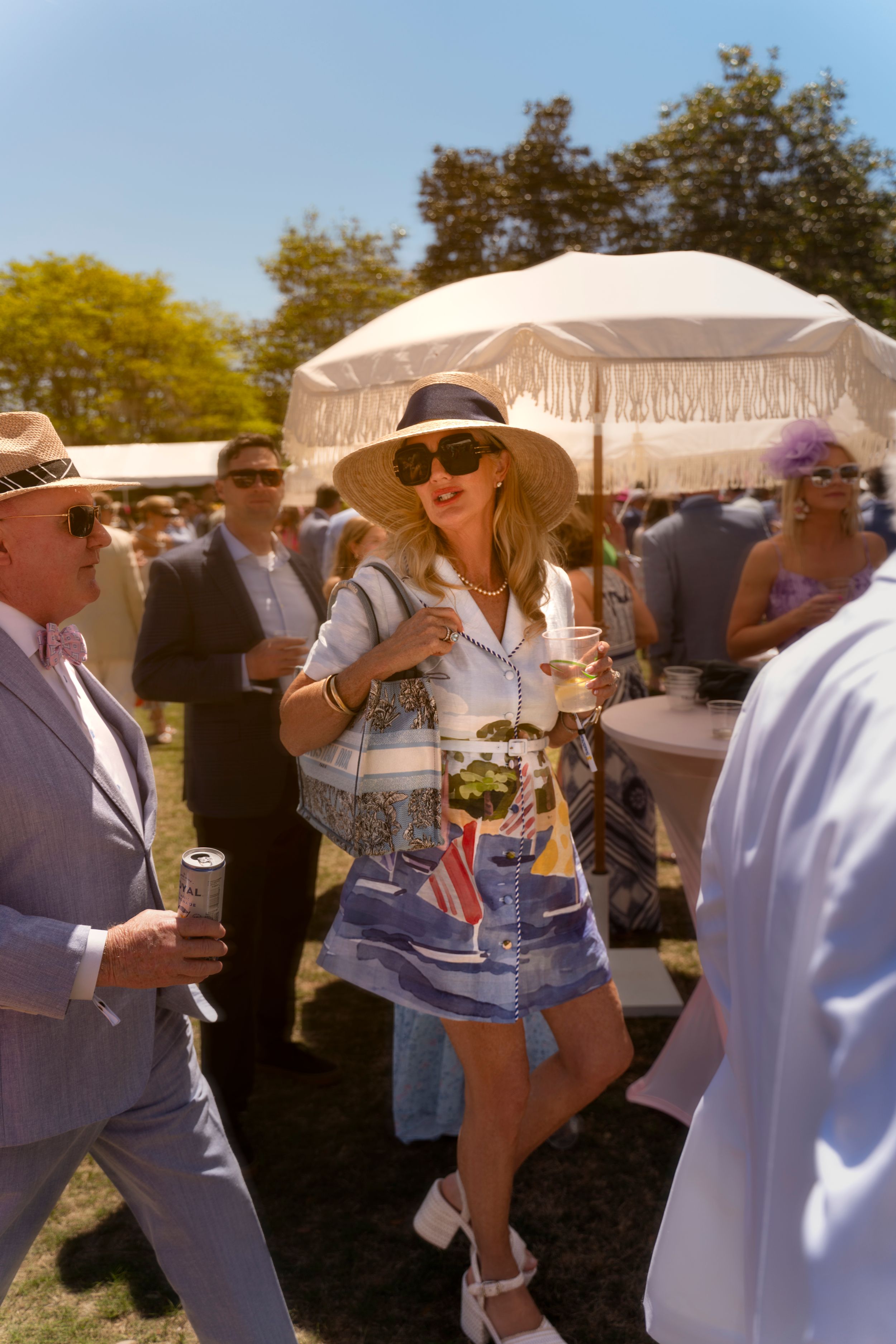 Woman in wide-brim hat, sunglasses, and denim skirt holding a drink at an outdoor summer garden party event.