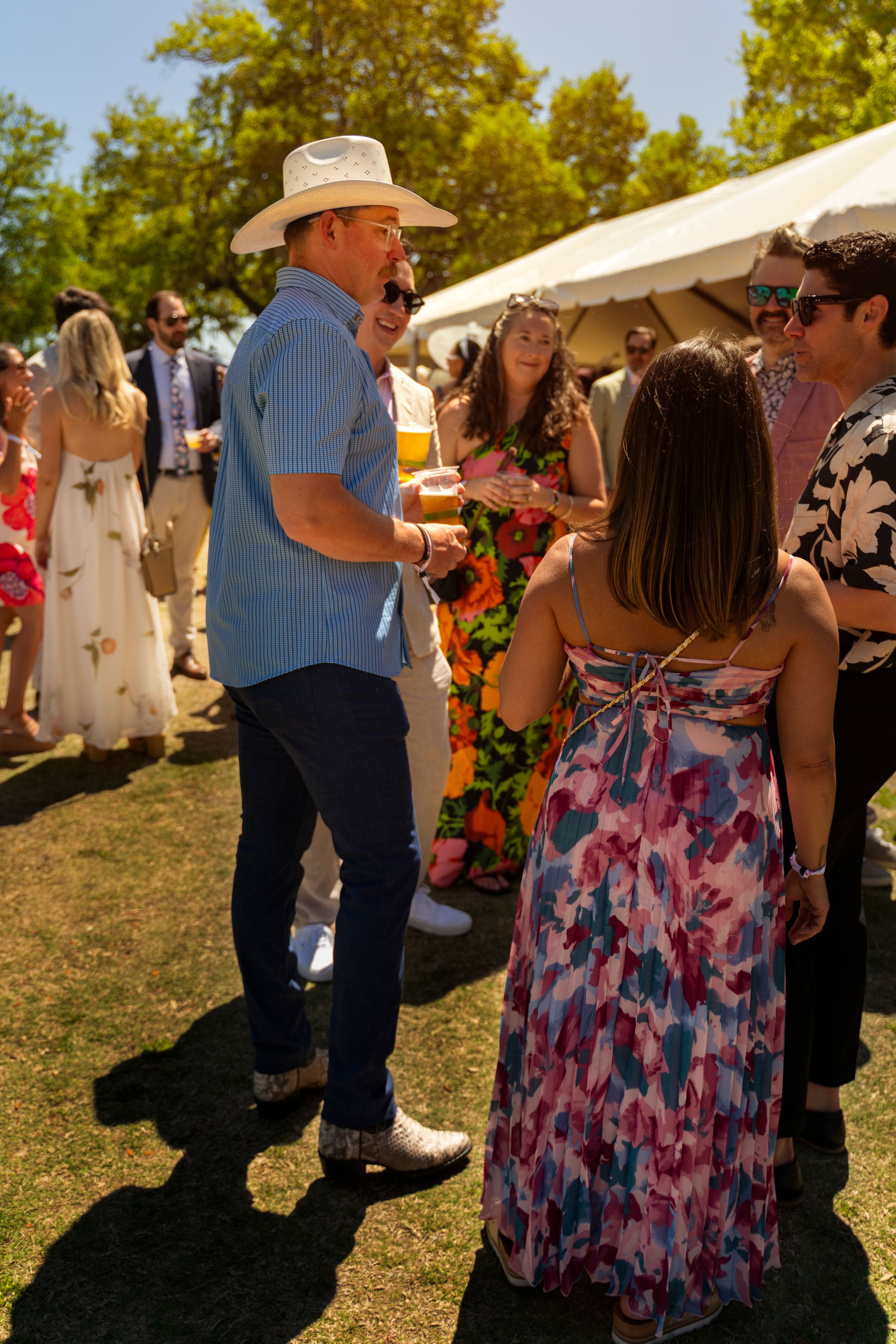 Guests in summer attire mingle outdoors at a garden party, man in cowboy hat and blue shirt holds a drink.
