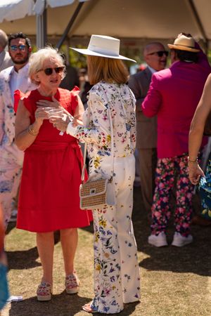 Two women in elegant floral outfits and hats socializing at outdoor summer event.