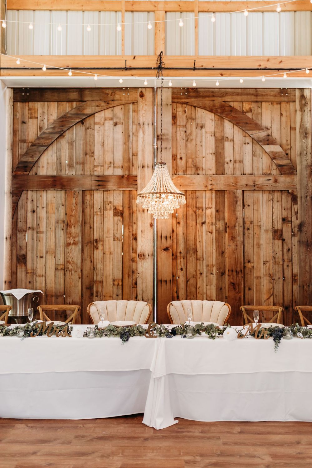 Rustic barn wedding reception with crystal chandelier, white draped sweetheart table, and floral garland against wooden doors.