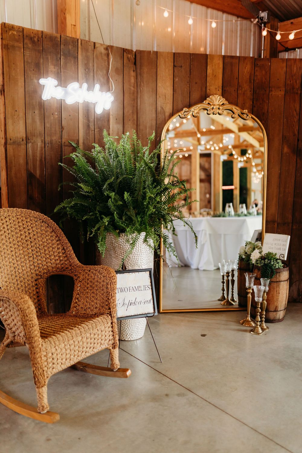 Rustic barn wedding decor with wicker rocking chair, gold mirror, ferns, candelabras, and string lights.