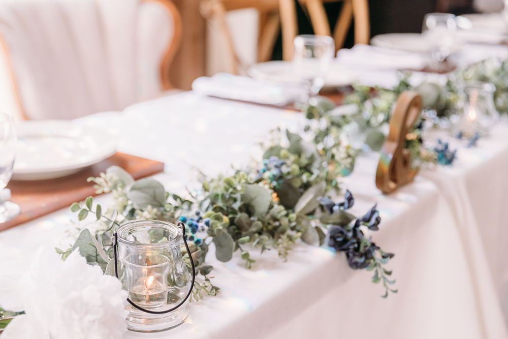 Elegant wedding table with eucalyptus garland, blue berries, and glass candle holders on white linen.