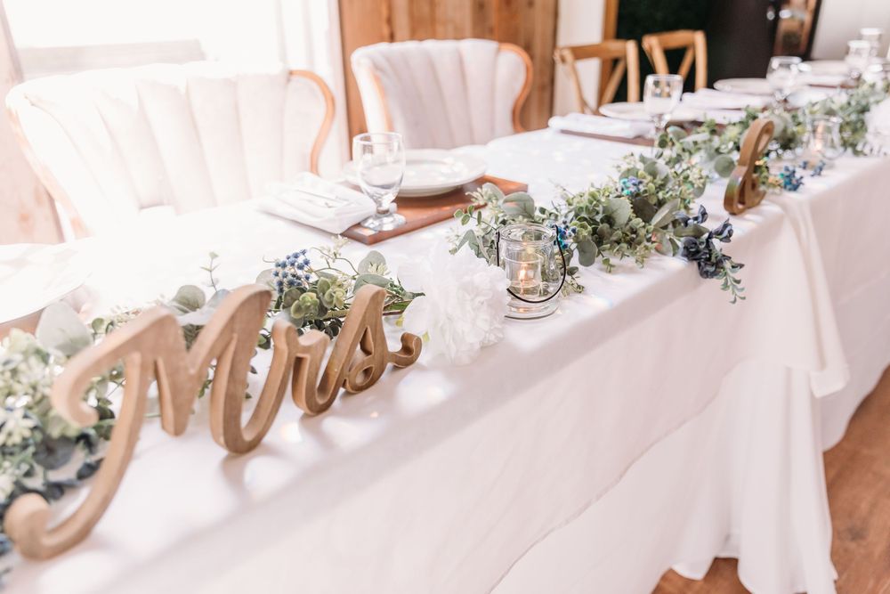 Elegant wedding head table with white linen, greenery garland, candles, and wooden Mr. & Mrs. signs.