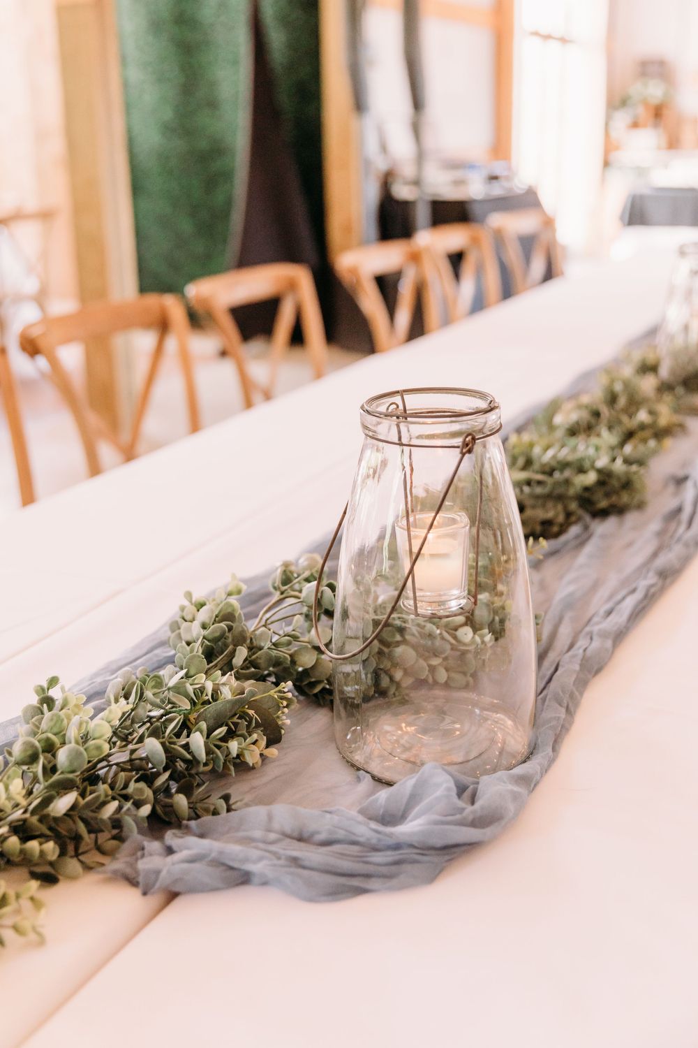 Glass lantern centerpiece with greenery garland and blue cheesecloth runner on white wedding reception table.