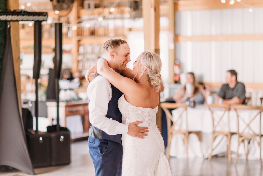 Bride and groom share their first dance in a rustic barn wedding venue with guests seated in the background.