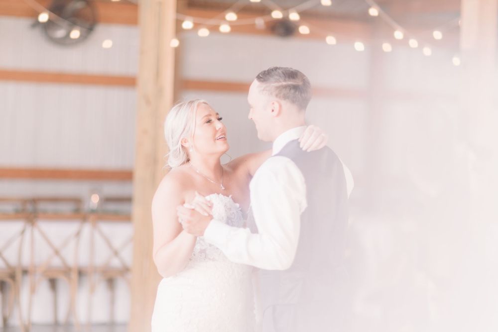 Bride and groom share their first dance in a warmly lit barn venue with string lights glowing softly above.