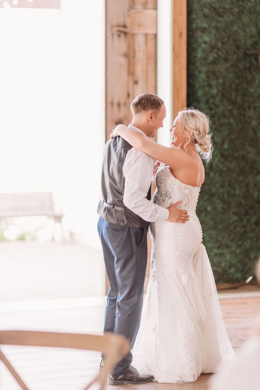 Bride and groom share their first dance in a rustic barn wedding venue with wooden doors and greenery wall backdrop.