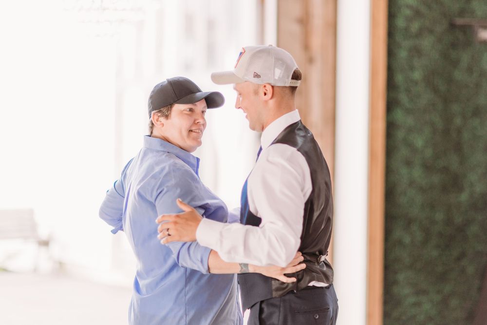 Two men in baseball caps share a warm embrace outdoors, smiling at each other in a bright, softly lit setting.