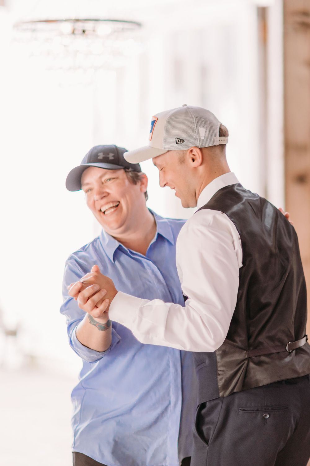 Two people in baseball caps share a joyful dance together at a wedding celebration.