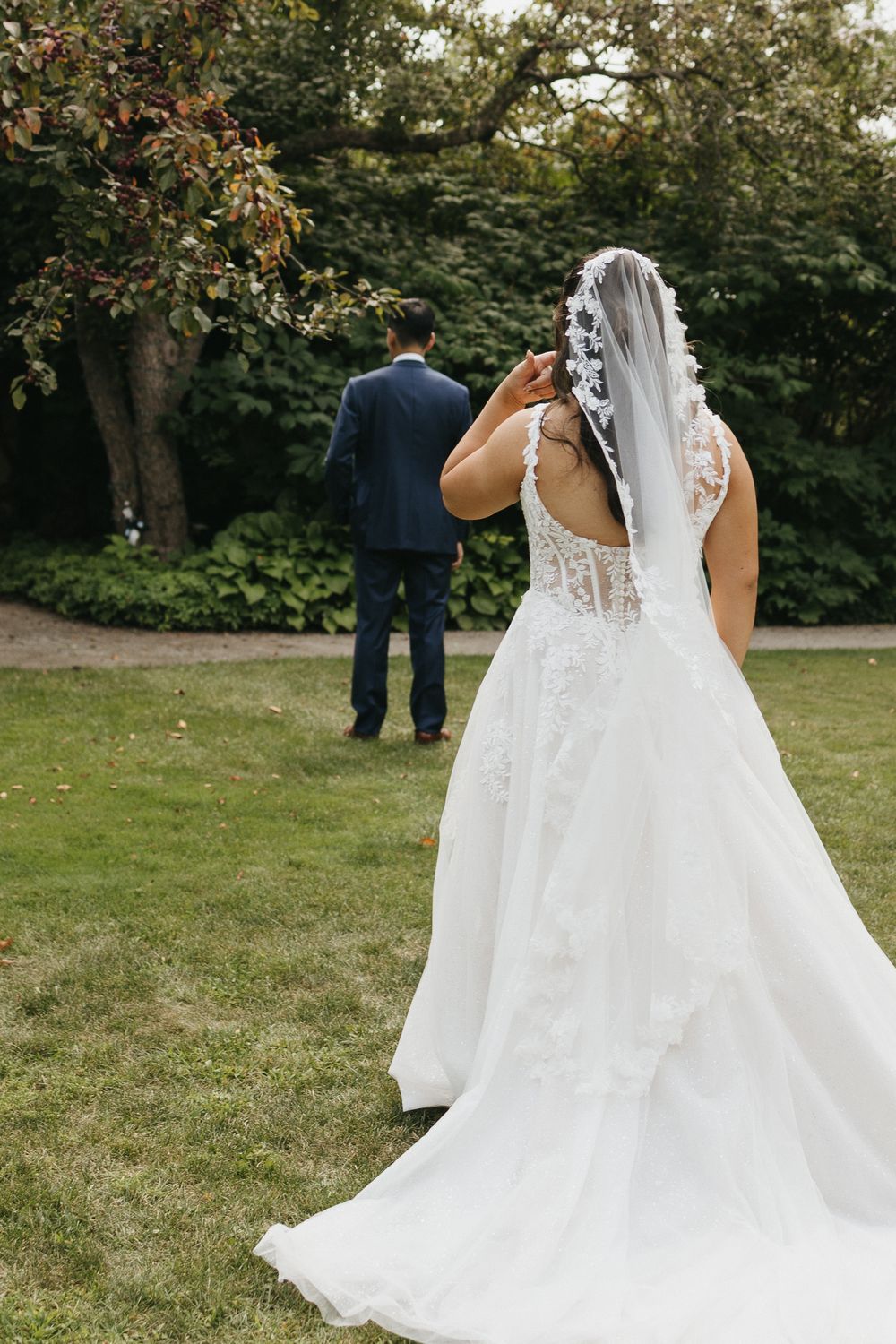 Bride in a lace ball gown and veil approaches groom in navy suit for a first look in a lush garden.
