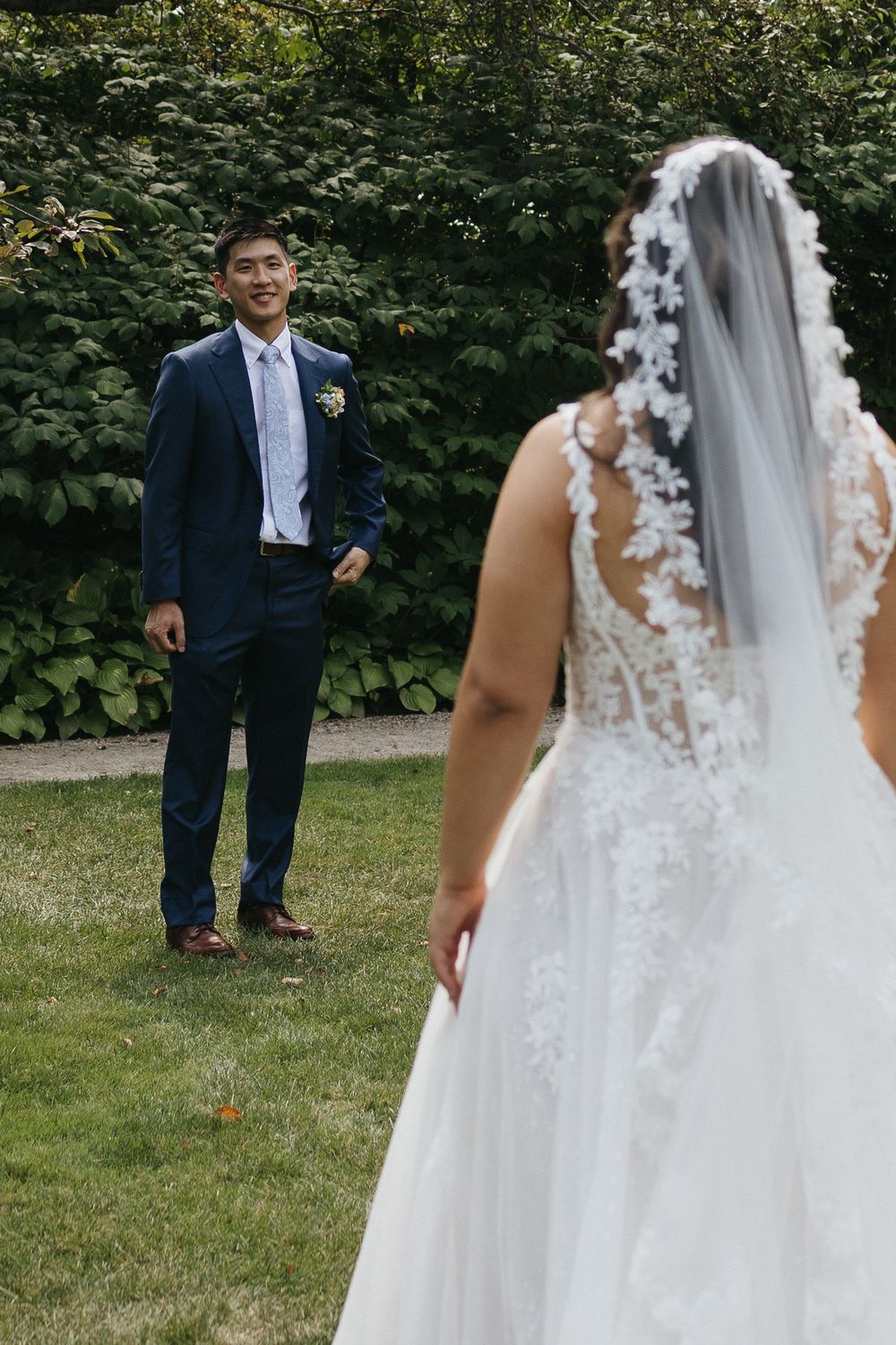 Groom in navy suit smiles at bride in lace wedding gown during outdoor first look on green lawn.