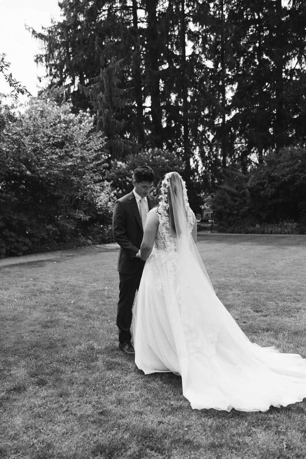 Black and white photo of bride and groom walking hand in hand across lush green lawn with tall trees.