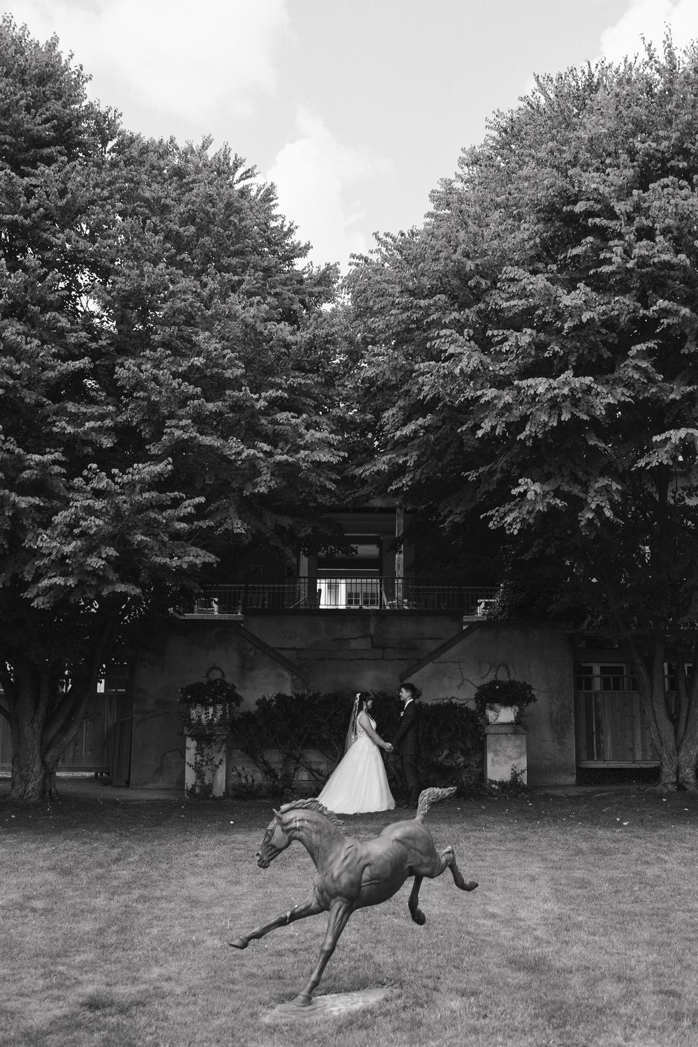 Black and white wedding portrait of bride and groom standing behind a horse sculpture in front of an estate.
