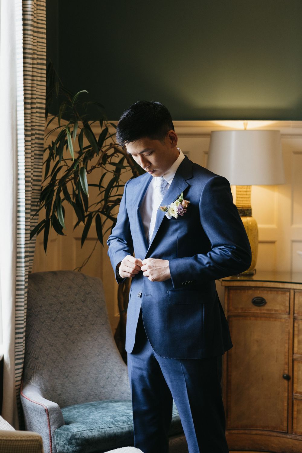Dapper groom in navy blue suit adjusting boutonniere while getting ready in elegant room.
