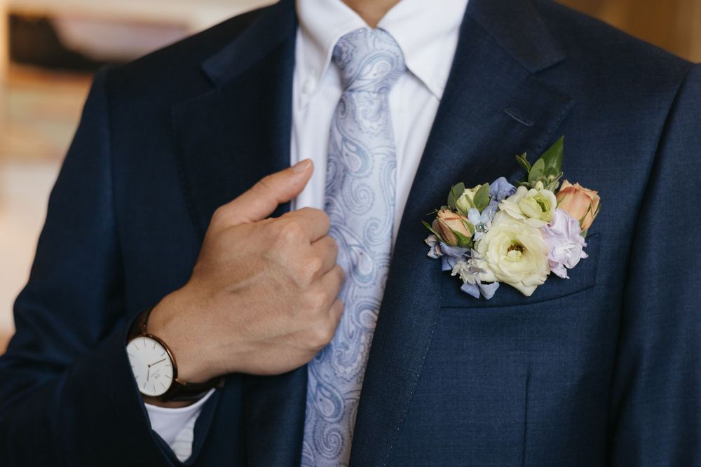 Groom in navy suit adjusts paisley tie with floral boutonniere featuring white ranunculus and blue flowers.
