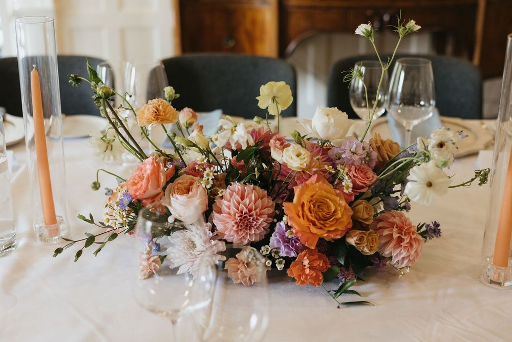 Elegant wedding table centerpiece with peach, orange, and white flowers including dahlias and roses with candles.