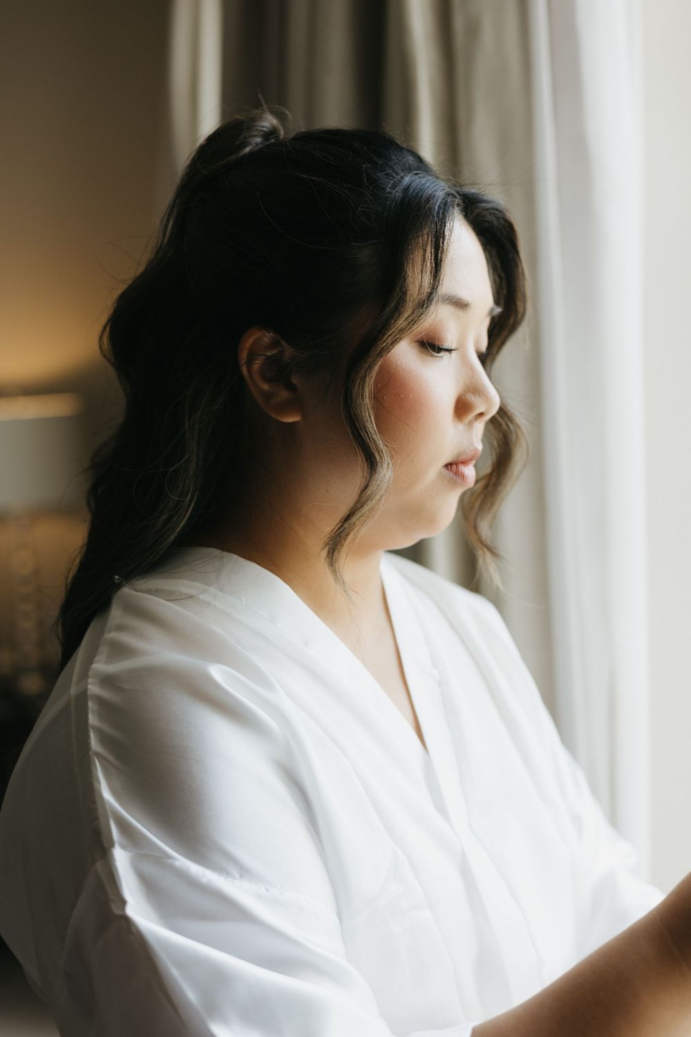 Bride in white robe with dark updo hairstyle gazing down by a window on her wedding morning.