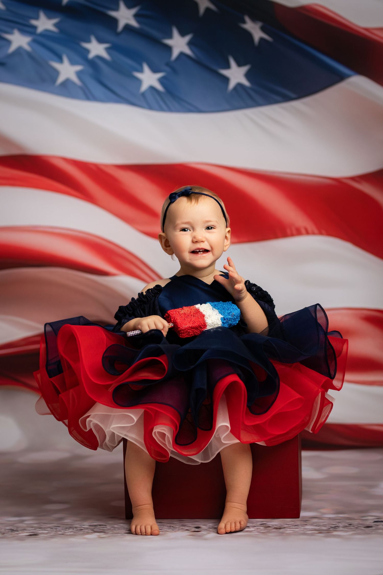 Adorable baby girl in patriotic red, white, and blue tutu dress sitting in front of American flag backdrop.