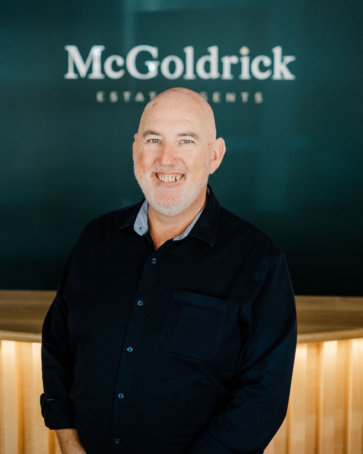 Smiling blonde woman in black lace shirt poses in front of McGoldrick Estate Agents teal office sign.