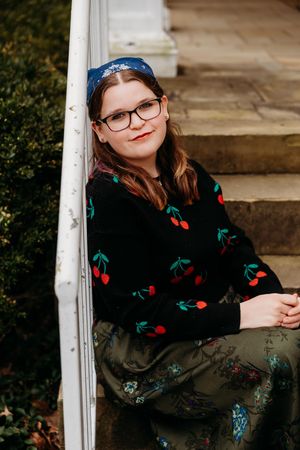 Young woman in cherry-print sweater and floral skirt sits on stone steps, wearing glasses and blue headband.