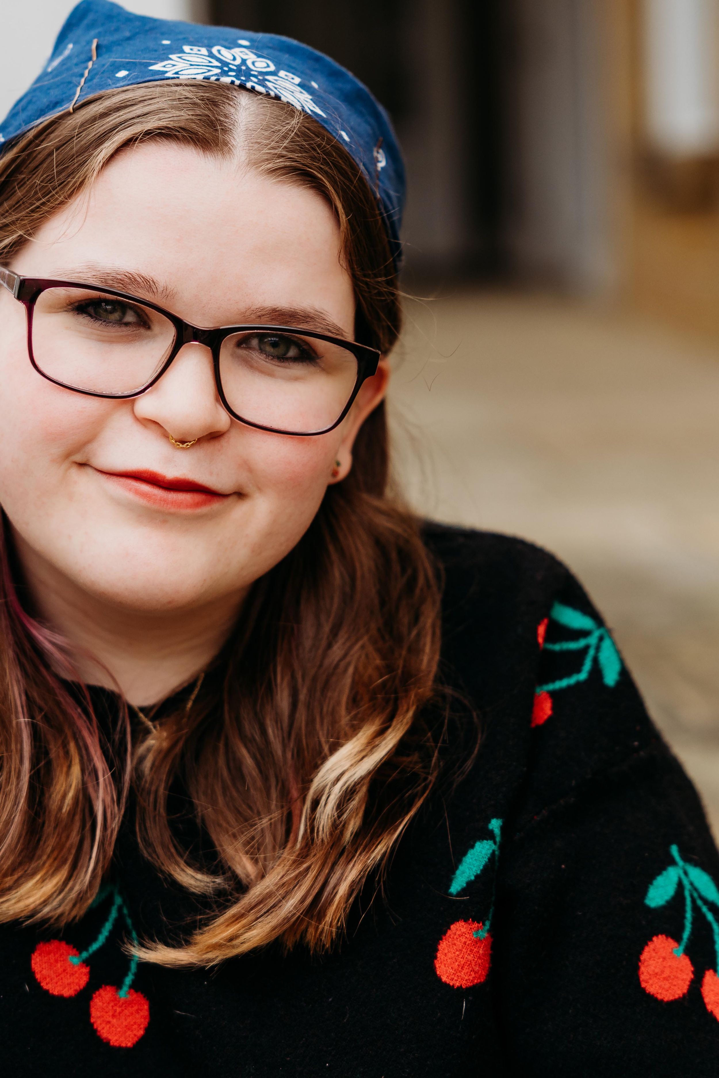 Young woman with glasses and blue headband smiles, wearing a black cherry-print sweater.
