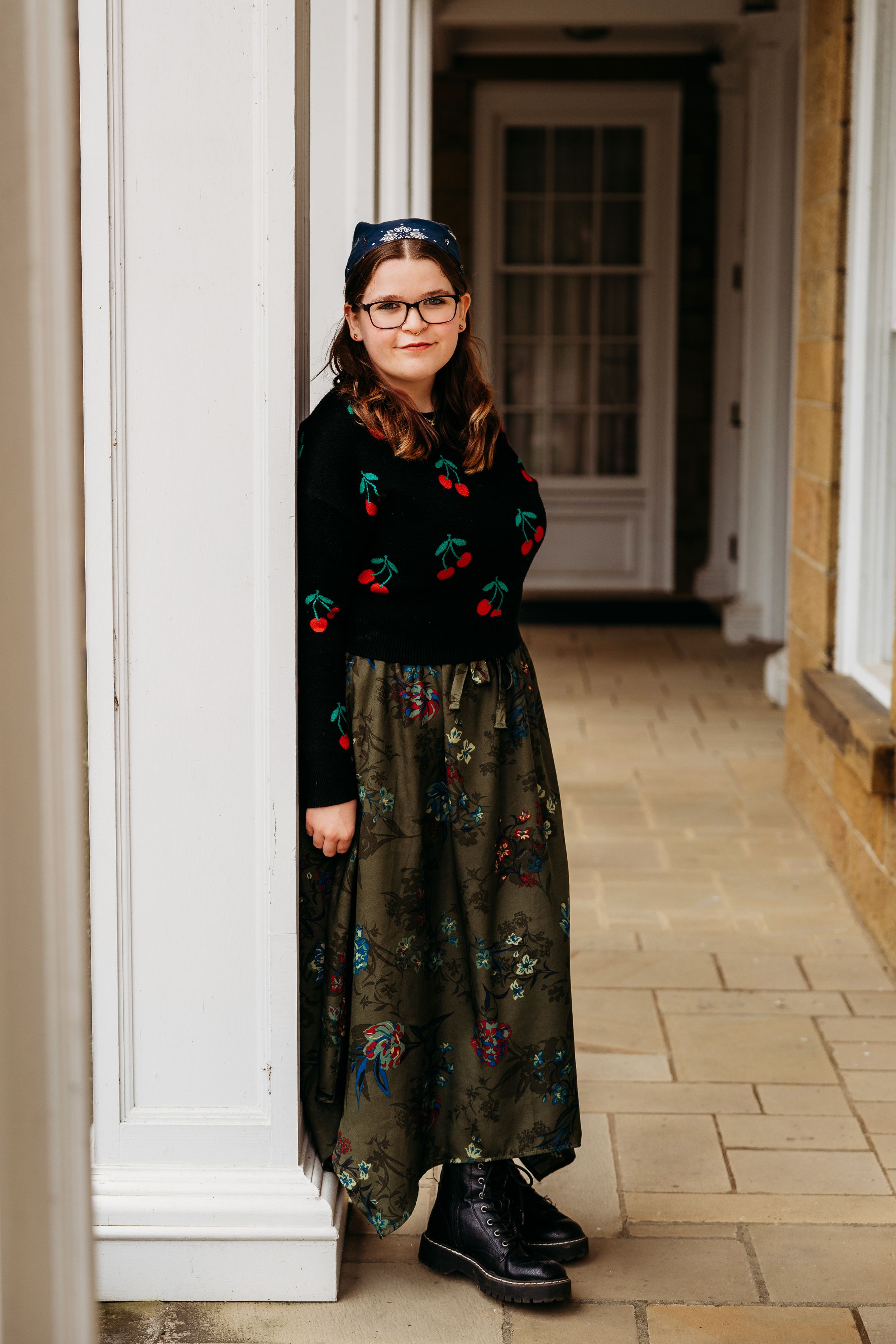 Young girl in cherry sweater and floral skirt leans against white column on a stone porch.