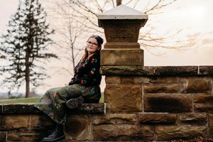 Young woman with glasses sits smiling on a stone wall outdoors, wearing a floral top and green pants in a park setting.