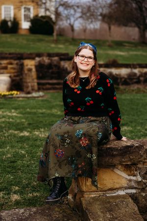 Young woman in cherry sweater and floral skirt sits smiling on stone wall in lush green garden.