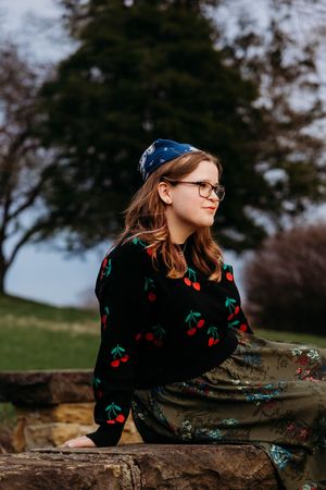 Young woman with glasses and blue headband sits outdoors wearing cherry print sweater and floral skirt in a park setting.