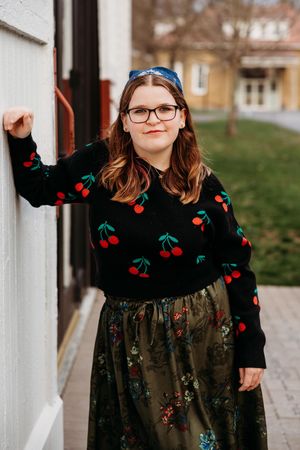 Young woman in cherry-print black sweater and floral skirt, wearing glasses, posing outdoors near a white wall.