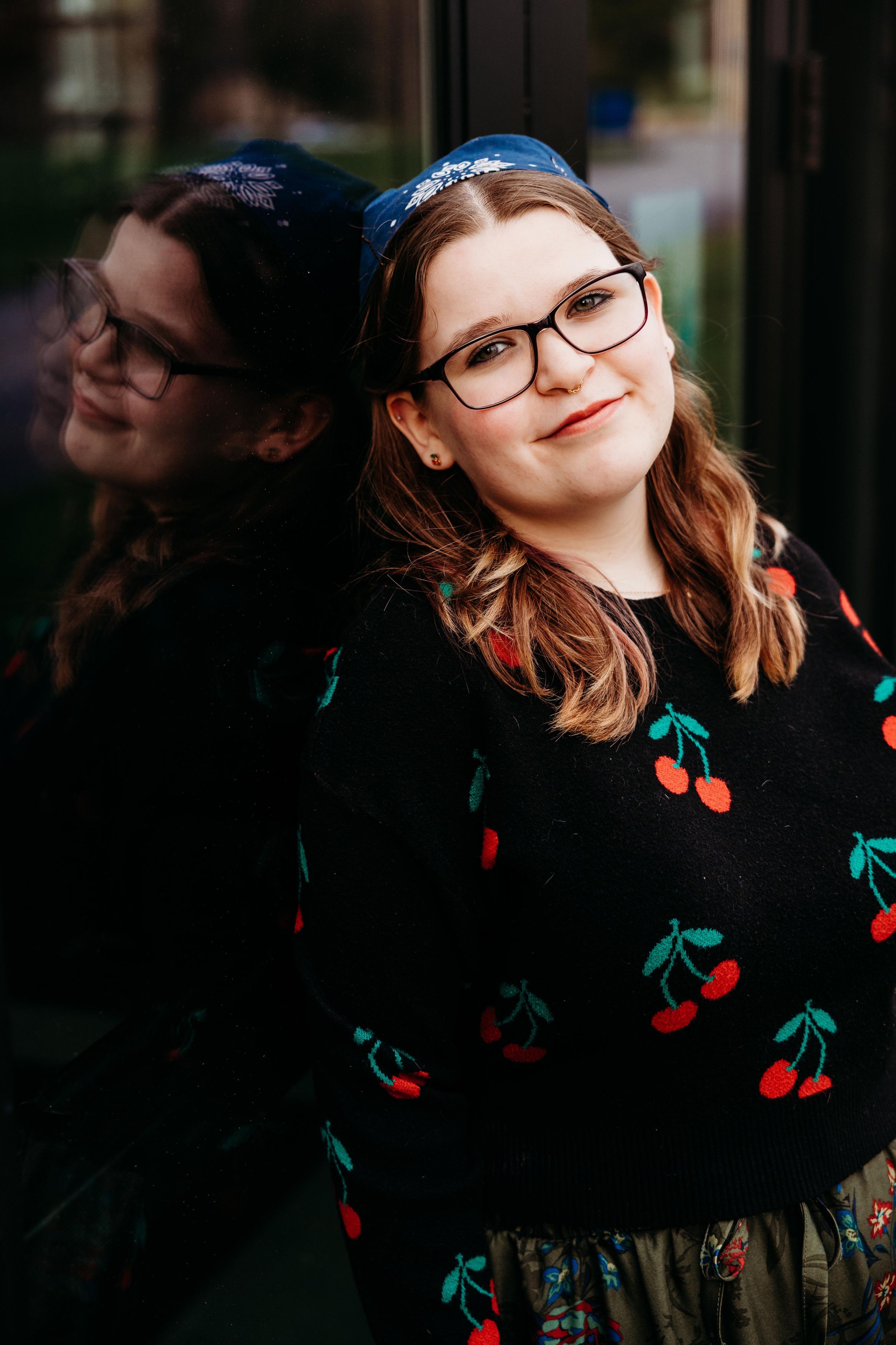 Young woman with glasses and blue headband wearing a cherry-print black sweater, leaning against a reflective glass surface.