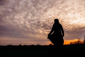 Silhouette of a woman in a flowing dress standing in a field against a dramatic sunset sky with clouds.