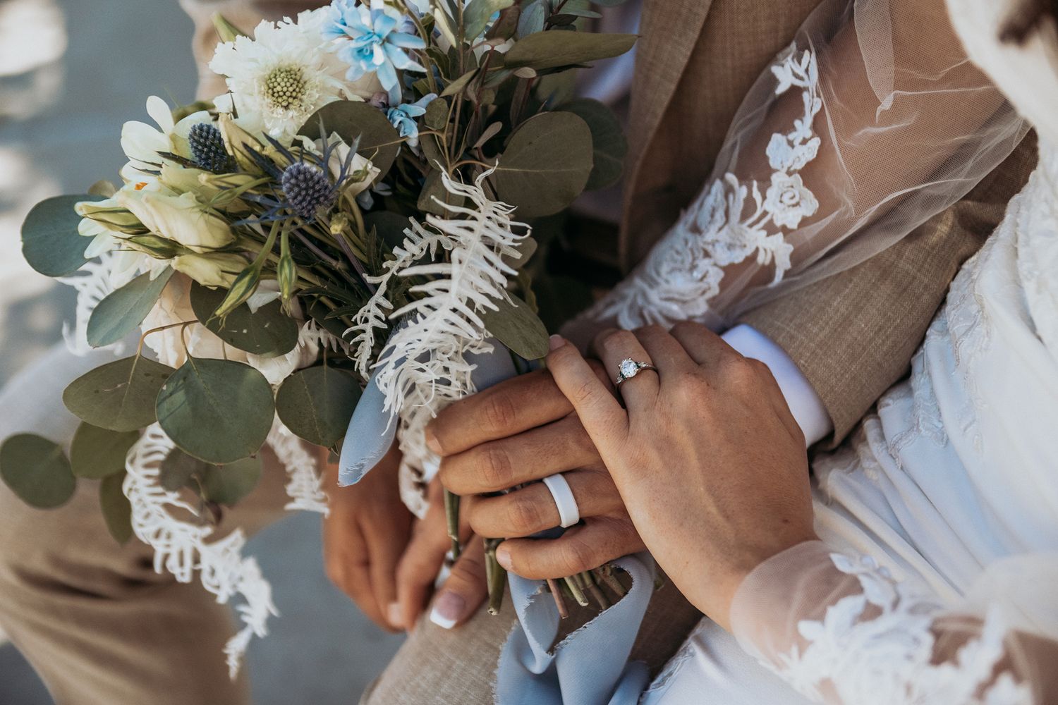 Newlywed couple holding hands with wedding rings and a rustic bouquet of white flowers and eucalyptus.