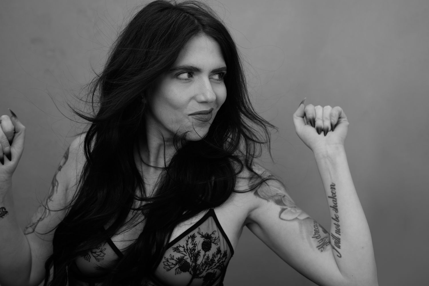 Black and white portrait of a woman with dark hair, tattoo on shoulder, posing gracefully against a gray background.