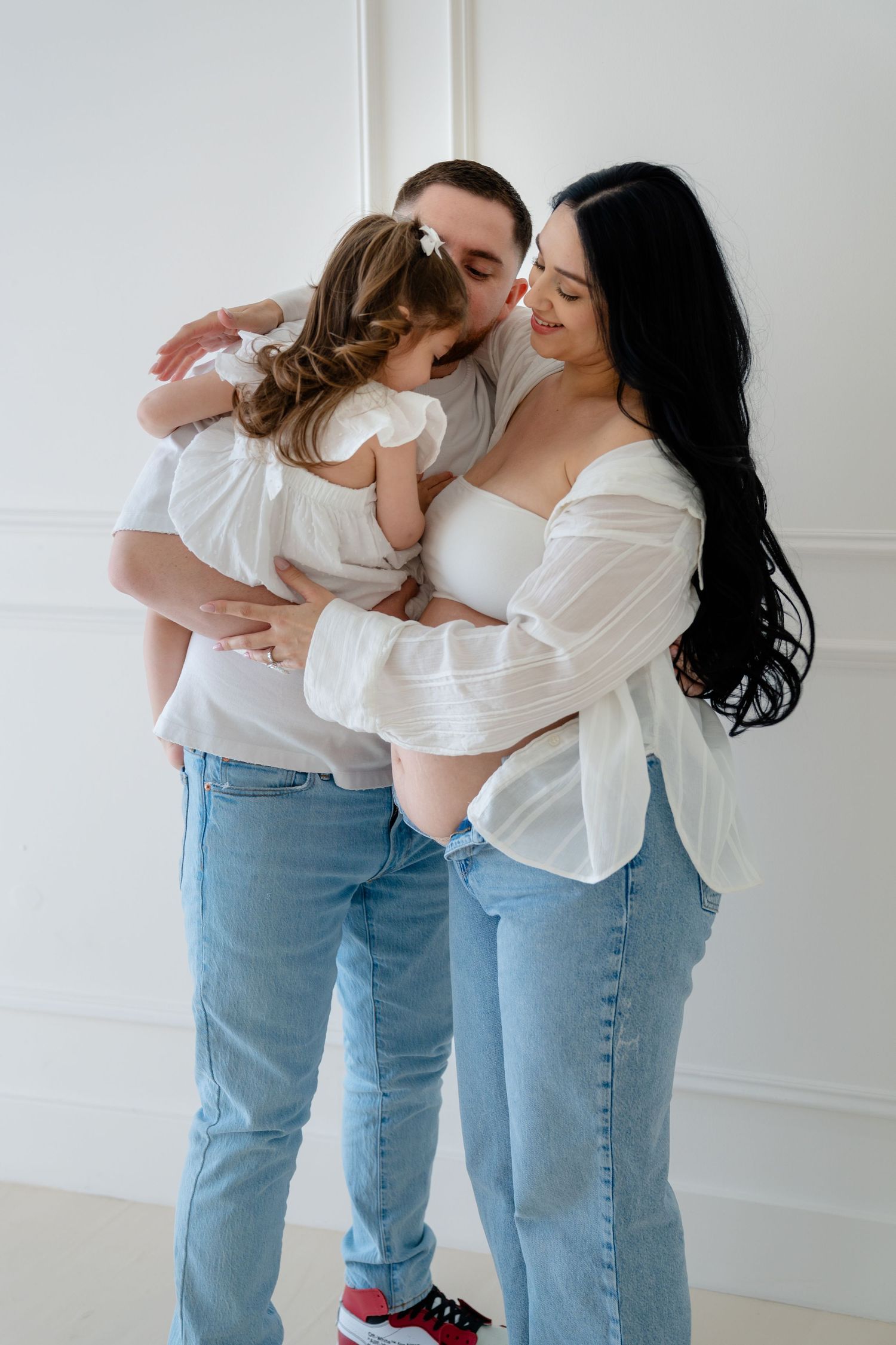 A happy family in white tops and jeans shares a tender embrace, with parents kissing their young daughter.