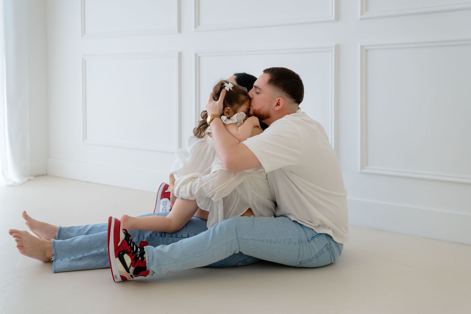 A couple sits on a white floor, sharing a kiss while a child in red sneakers and jeans leans between them.