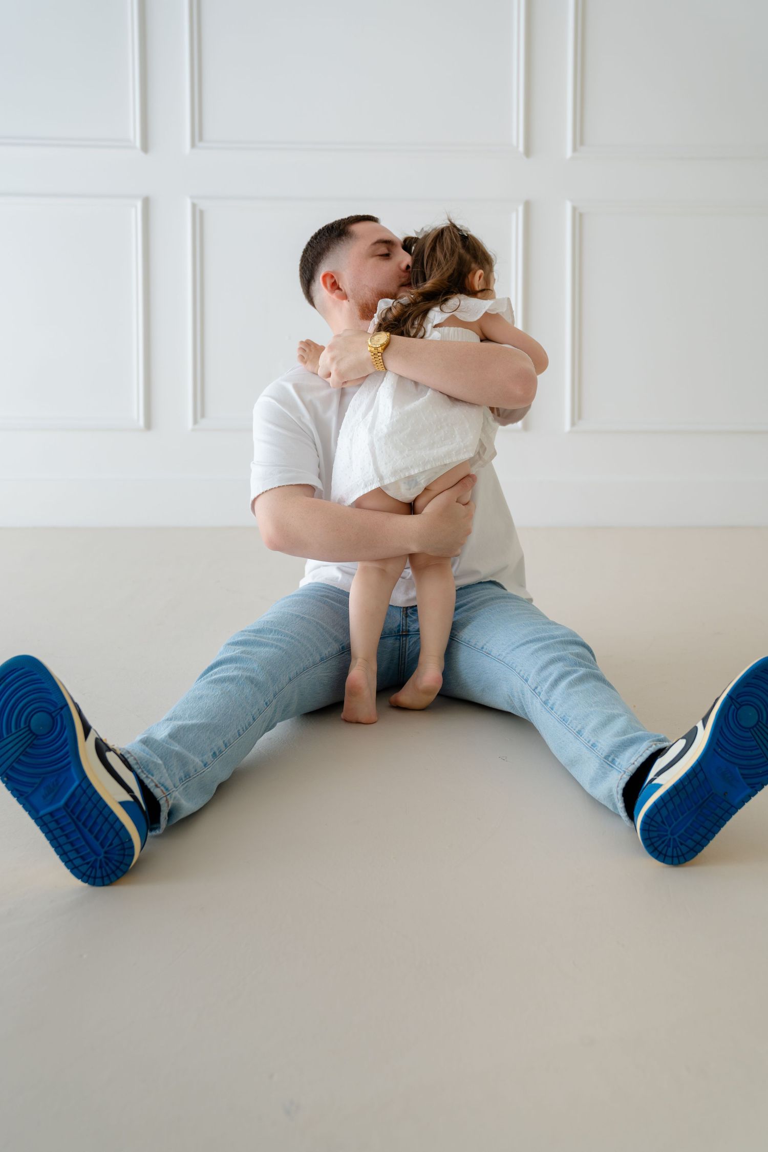 A father sitting on the floor hugging and kissing his young toddler daughter in a bright white room.