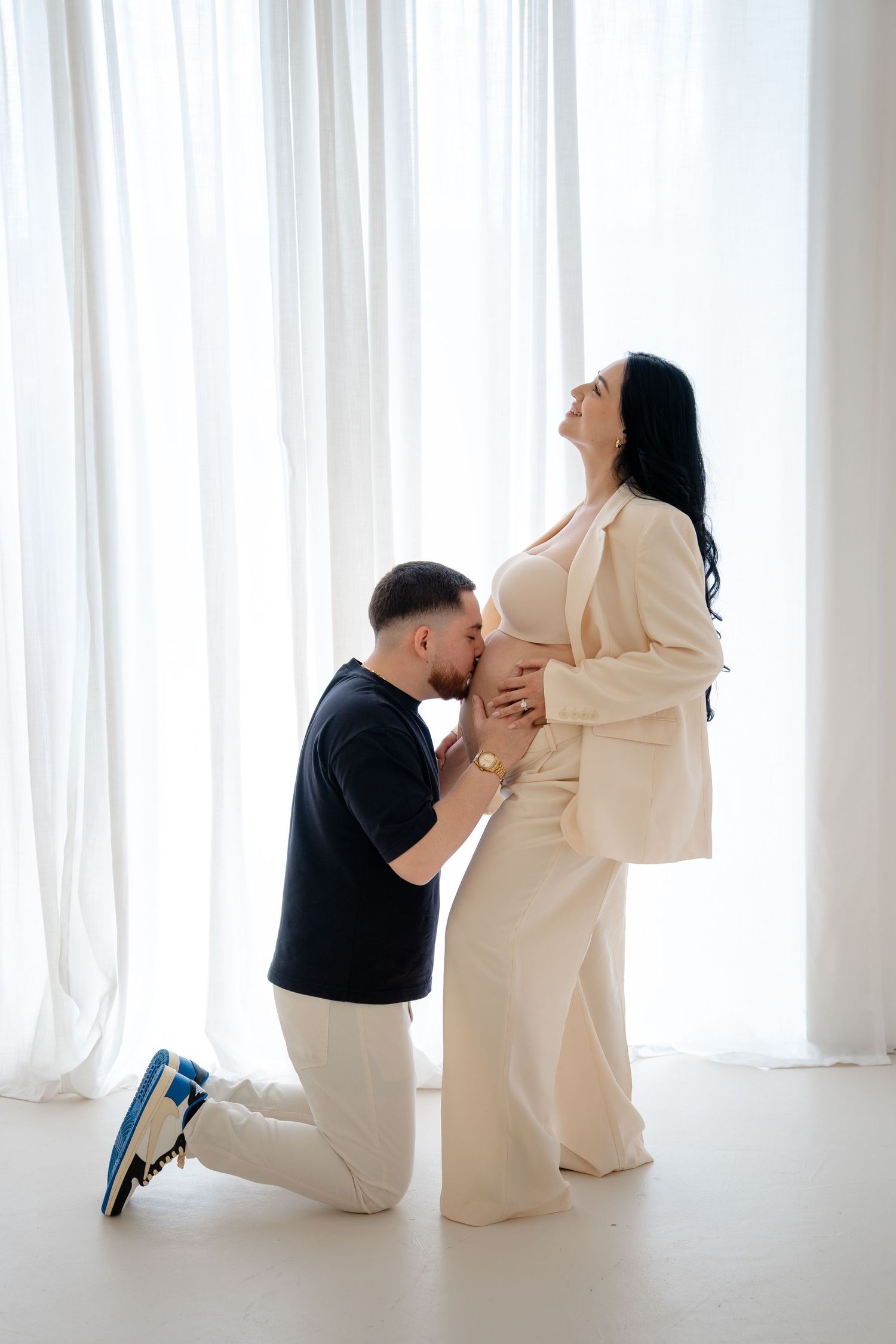 A man kneeling and kissing a pregnant woman's belly while she smiles, both dressed in white outfits.