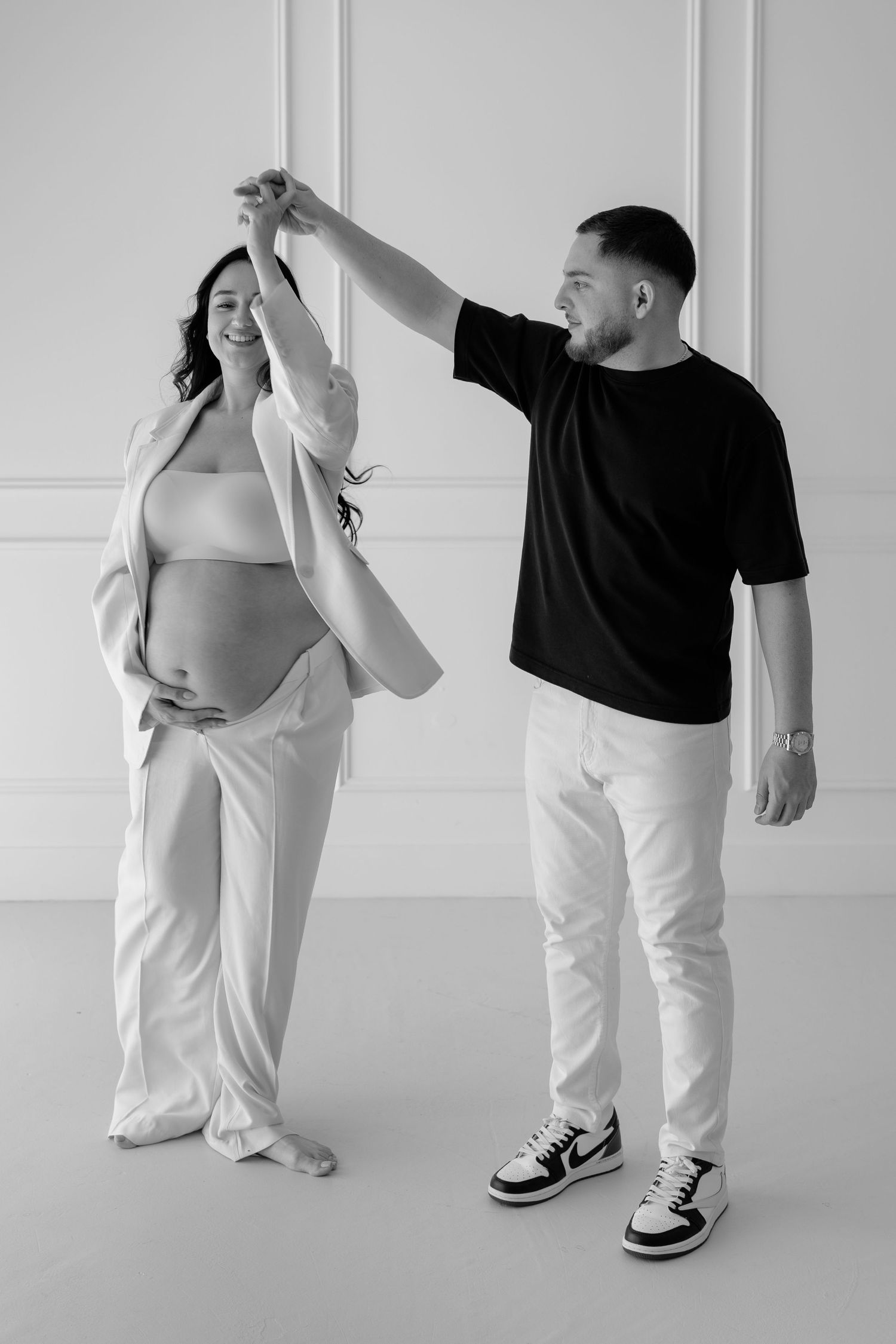Black and white photo of a happy pregnant couple dancing together in a bright white studio setting.