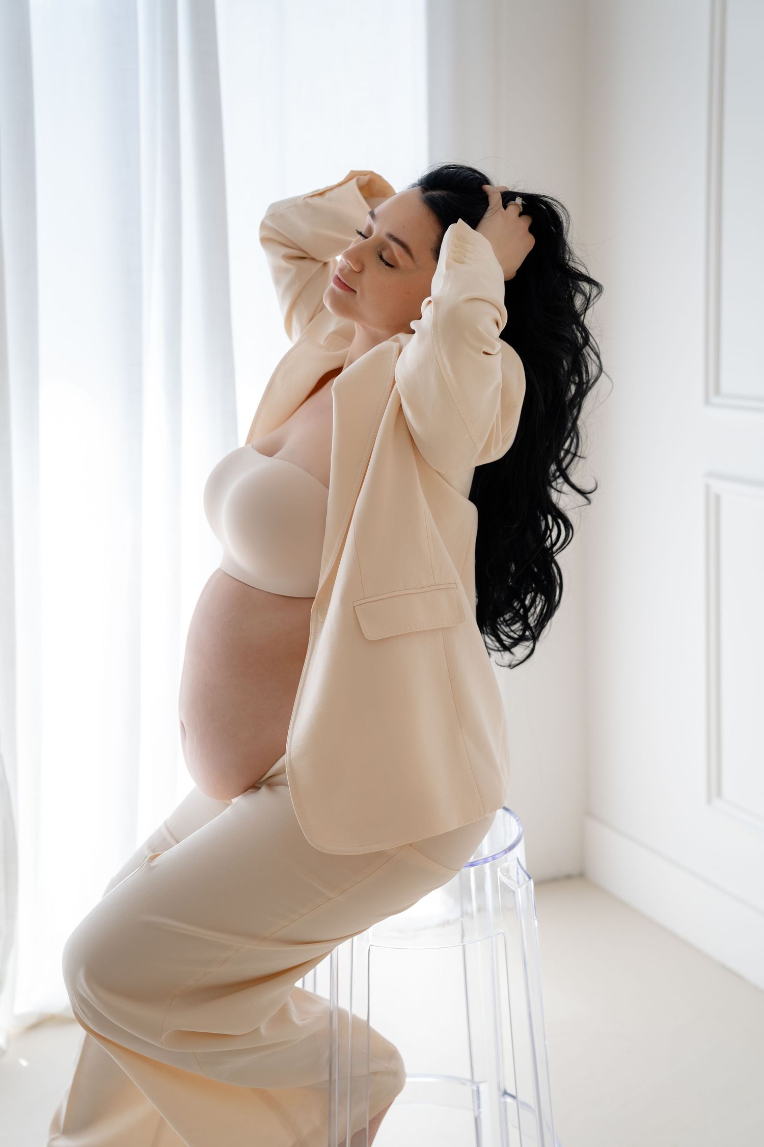 Pregnant woman in beige blazer and lingerie sits gracefully on clear chair, posing with hands in dark wavy hair.