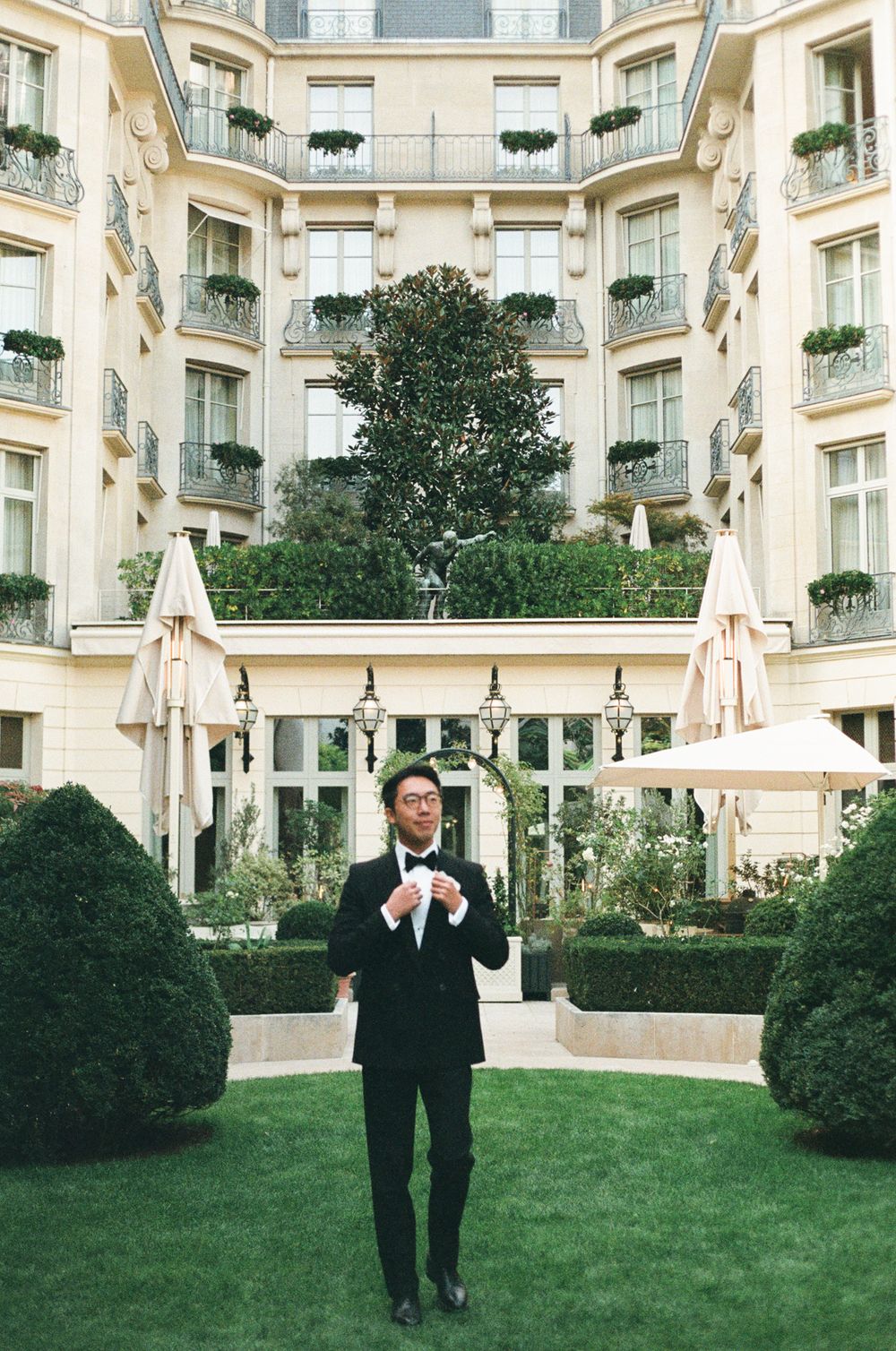 A man in a black tuxedo adjusts his bow tie in a lush hotel courtyard garden in Paris.