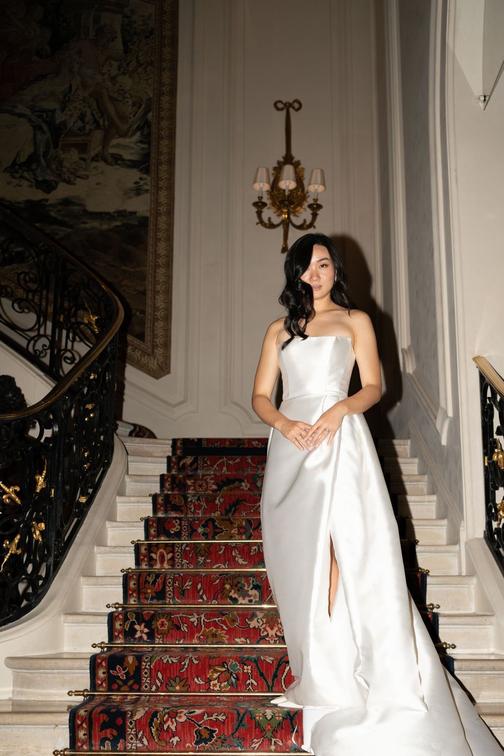Bride in strapless white satin gown with slit poses on ornate staircase with red carpet and gold chandelier.