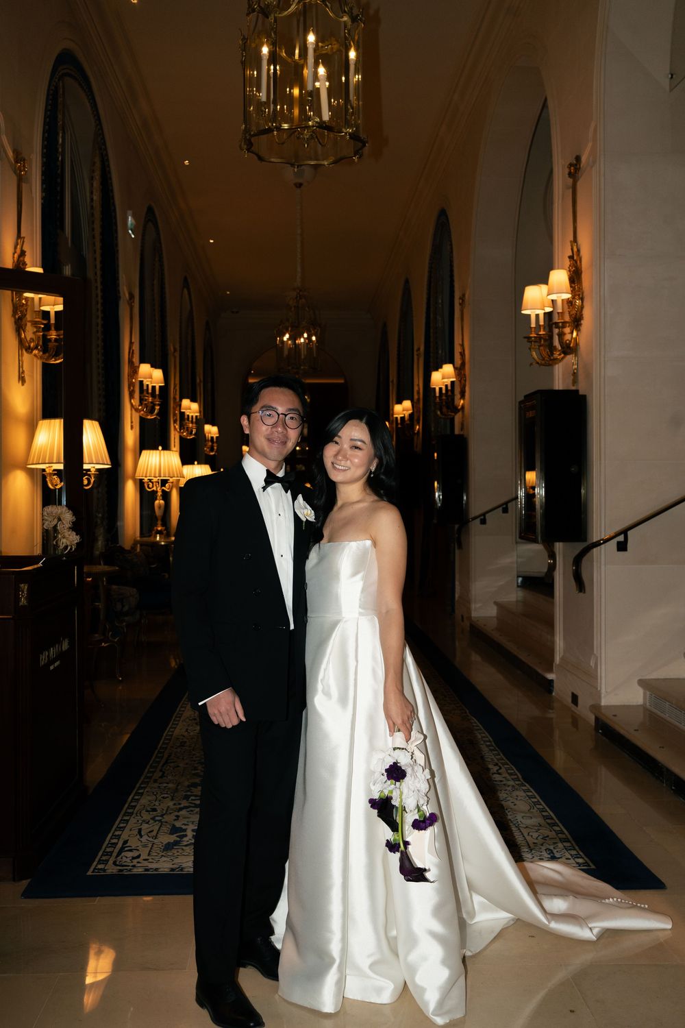 Bride and groom posing in elegant chandelier-lit hotel hallway, bride in strapless white gown holding purple bouquet.